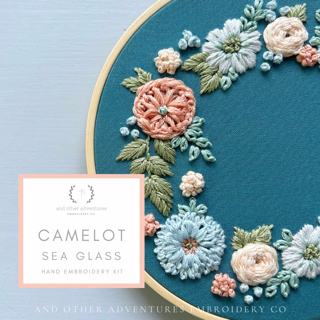 Embroidery hoop with floral embroidery design on a teal fabric, featuring the brand 'Camelot Sea Glass' and 'And Other Adventures Embroidery Co'.