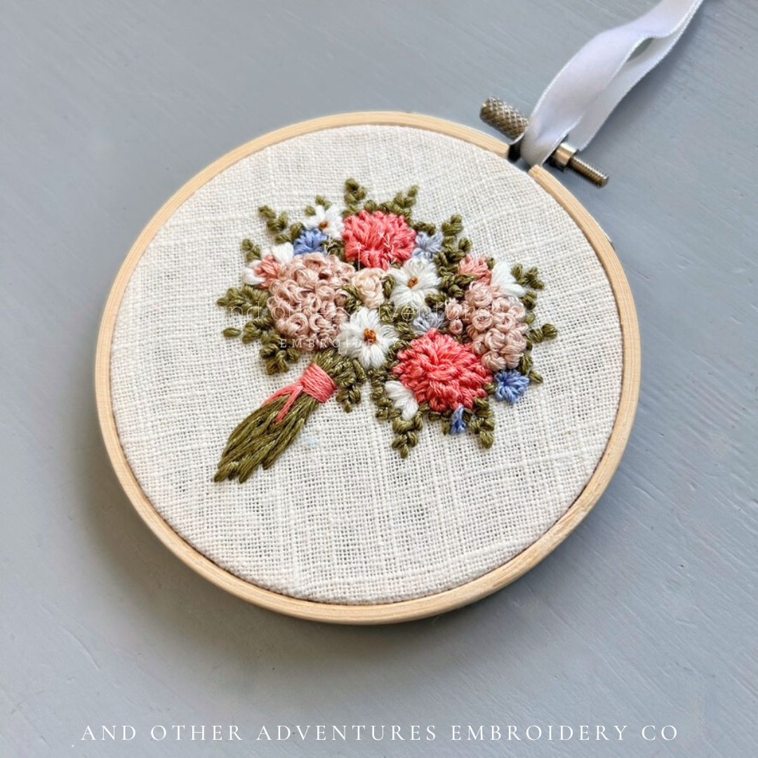 Embroidery hoop with floral design on a gray background, featuring &#39;And Other Adventures Embroidery Co&#39; branding.
