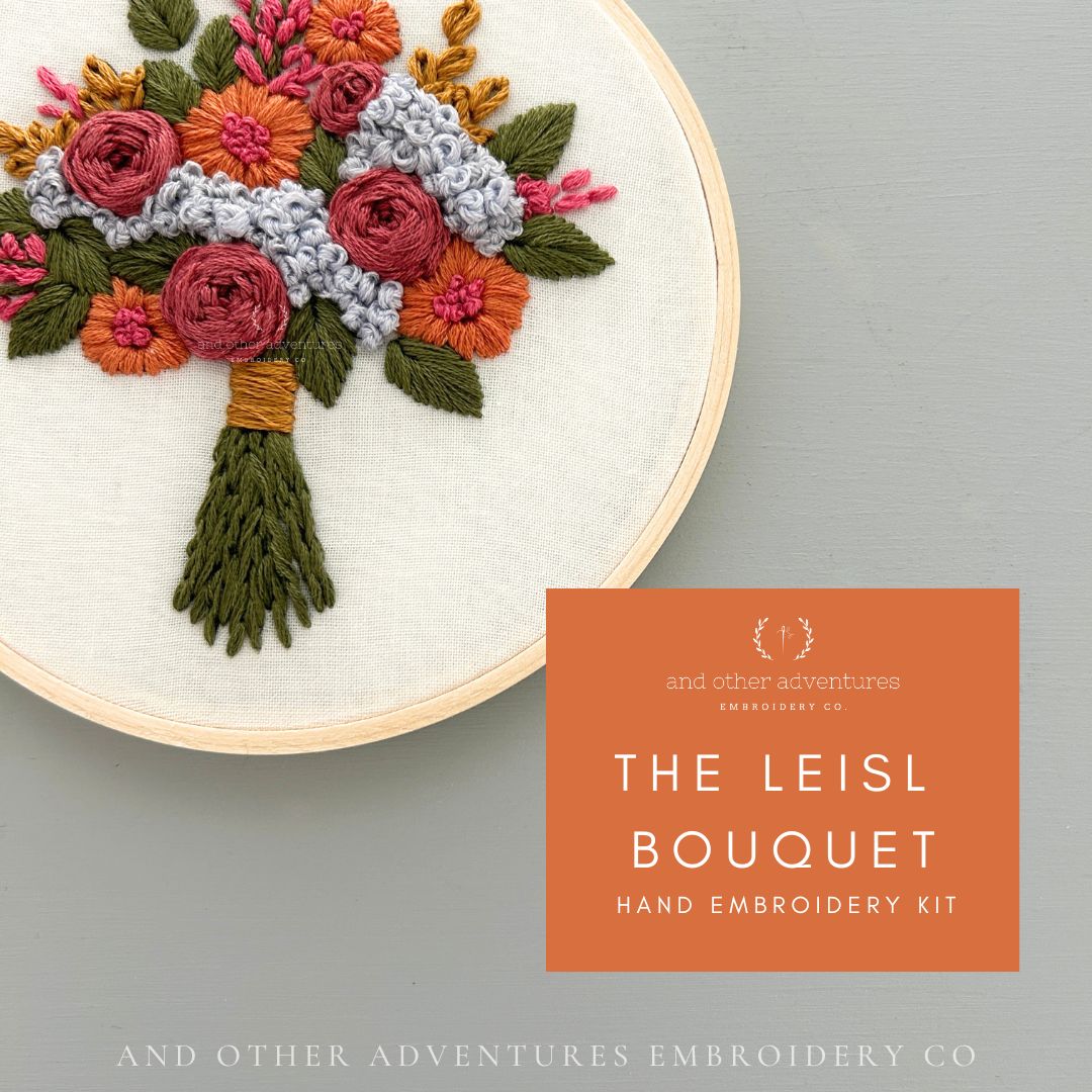 A hand embroidery kit showing a completed floral bouquet embroidery in a wooden hoop on a light gray background, with an orange label reading 'THE LEISL BOUQUET HAND EMBROIDERY KIT' and 'and other adventures embroidery co'.