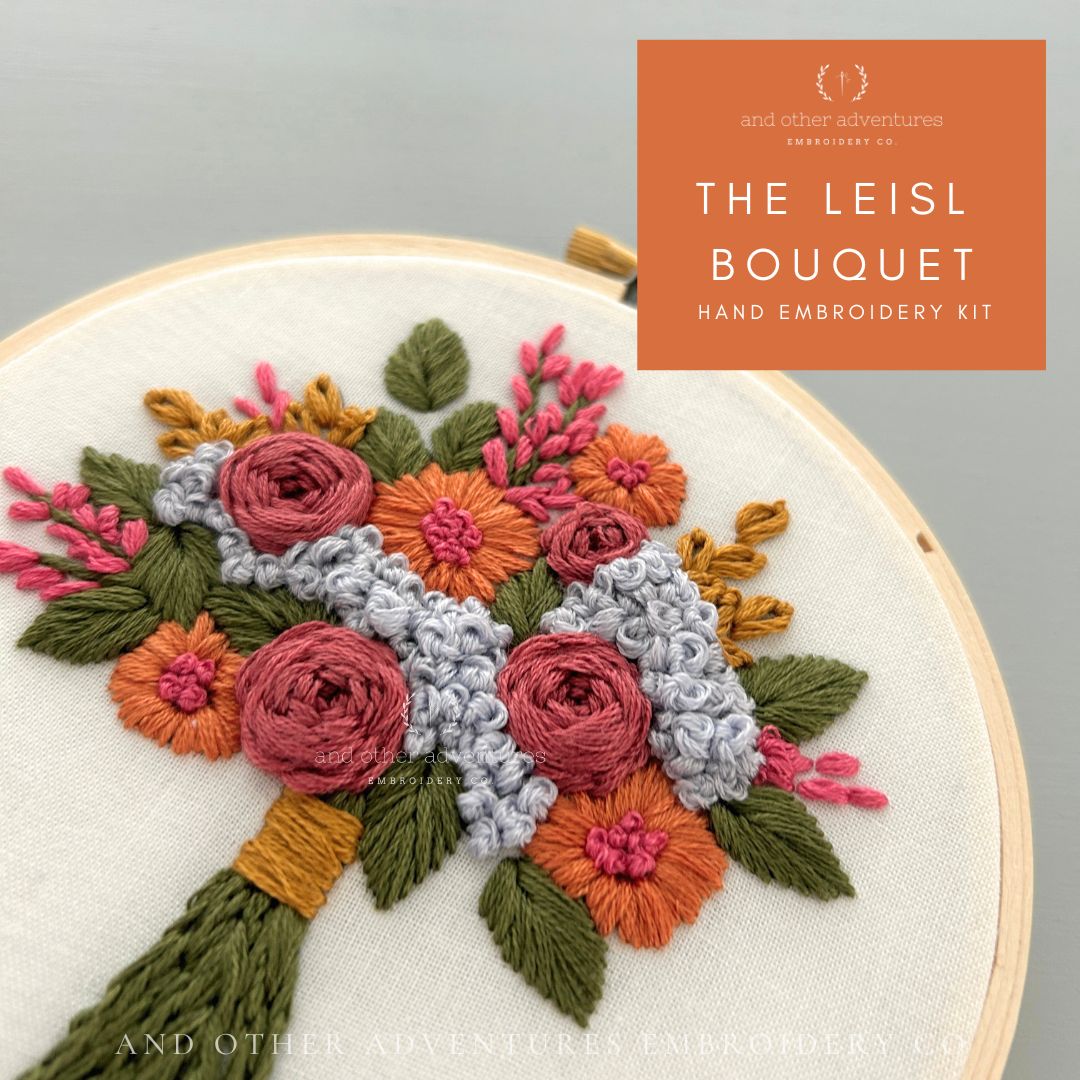 A close-up of a hand embroidery bouquet design inside a wooden hoop, with an orange label reading 'THE LEISL BOUQUET HAND EMBROIDERY KIT' and 'and other adventures embroidery co.' The bouquet features pink, orange, green, and light gray embroidered flowers and leaves on white fabric.  Designed, stitched, and sold by And Other Adventures Embroidery Co