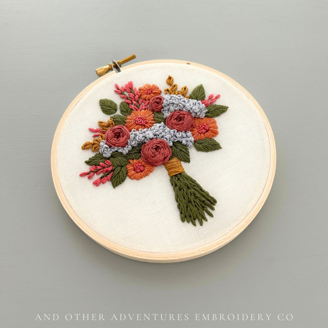 DIY hand embroidery hoop art featuring a hand stitched bouquet of flowers in shades of mauve, orange, green, pink and icy blue. Designed, stitched, and sold by And Other Adventures Embroidery Co