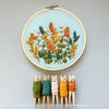 Embroidery hoop with floral pattern on a light mint green background, accompanied by colorful embroidery floss on wooden clothespins. And Other Adventures Embroidery Co